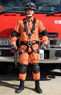 救助工作車の前にオレンジ色の救助服を着て、肘当てと膝当て、安全帯を着けた救助隊員が立っている写真