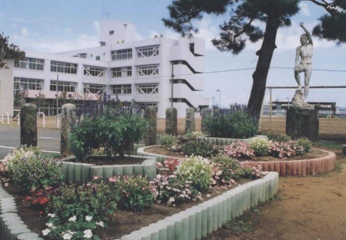 学校の2つの花壇に赤や紫の花が咲いている横に松の木と左手を上に上げている裸の銅像があり、奥に4階建ての校舎が写っている写真
