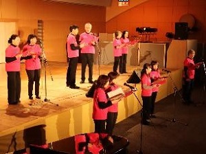 ピンク色の揃いのTシャツを着て、男女10数人がステージで歌っている様子の写真