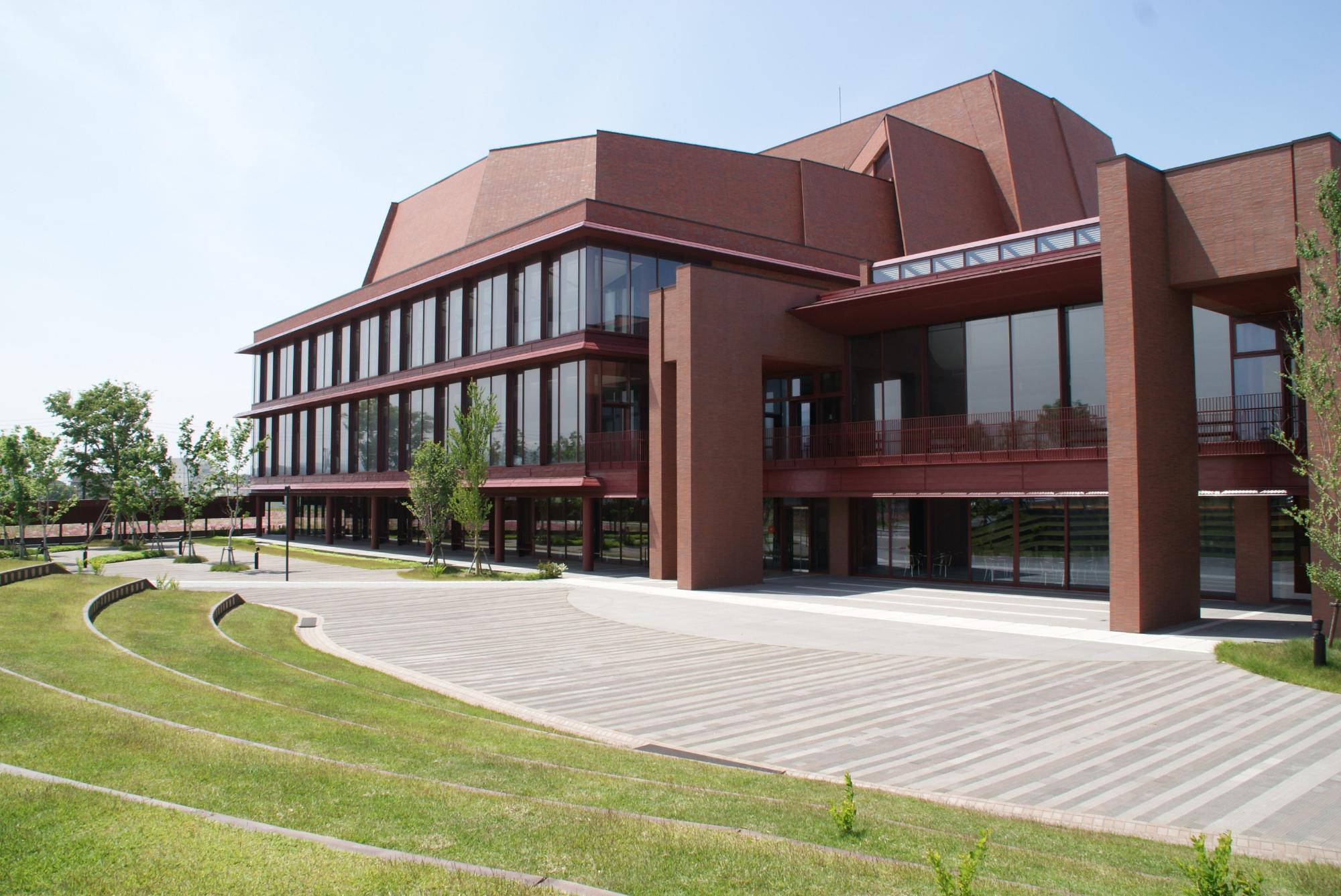 写真：手前に芝生が植えられた階段状の観客席があり、インターロッキングの発表ステージがあります。その奥にはアルフォーレの建物があります