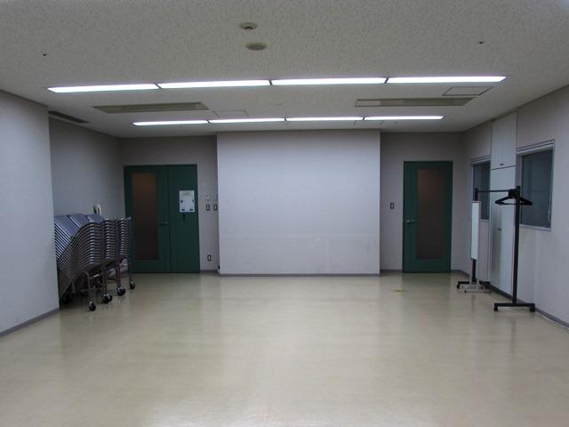 白い壁・床・天井の部屋の奥に、緑色の扉が2枚あり、左側の扉の前にパイプイスが積んである写真