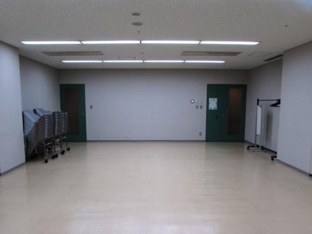 白い壁・床・天井の部屋の奥に、緑色の出入り口が2つあり、左手の出入り口前にはパイプイスが積まれている写真