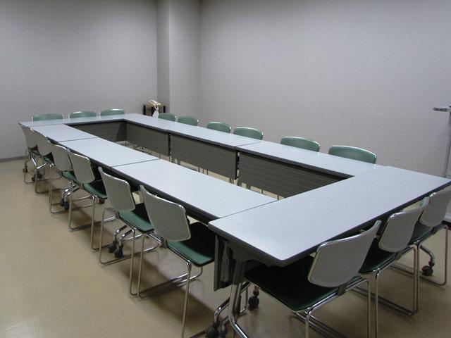 白い壁と床の室内に、白い長テーブルと座面が緑色のイスが並んでいる写真
