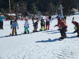 写真：参加者がスノーボードをつけ、少し膝を曲げた基本の姿勢を教えてもらっている様子です