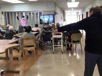 写真：入居者が集まりラジオ体操をする様子。映像に合わせてのびの運動をしています