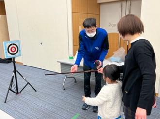 写真：スポーツ推進委員が子どもにスポーツ吹き矢を渡している様子。的が近くに写っています