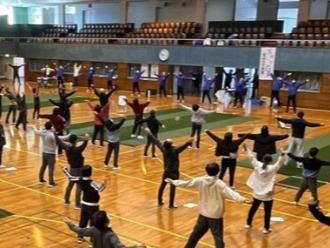 写真：スポーツ推進委員が模範となりラジオ体操をしています。参加者の大勢が後ろそりをしています。