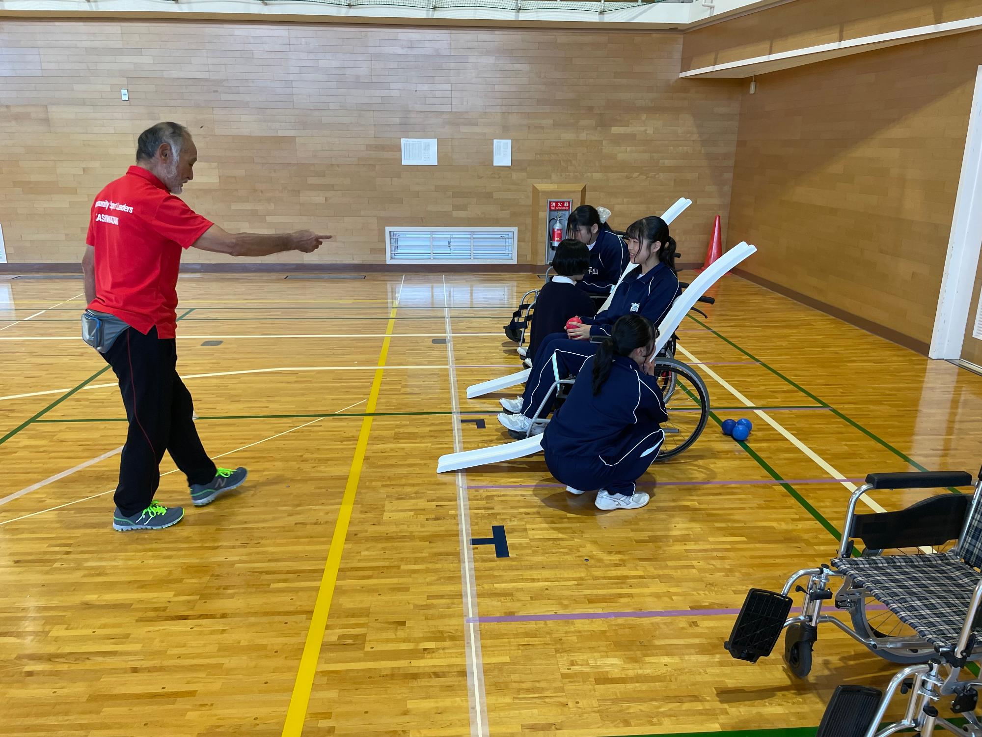 写真：ボッチャ体験の様子。ボールを転がすための用具がついた車椅子に座った生徒に、スポーツ推進員が使い方を指導しているところ