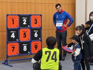 写真：小さな子供がスポンジでできたディスクを投球する様子を、大人や子供が周りで見守っています