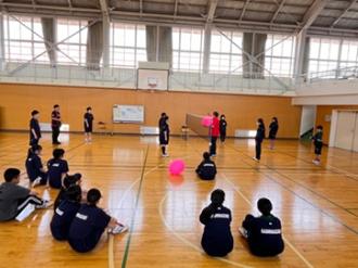 写真：赤いポロシャツを着たスポーツ推進委員が、バドミントンコートでふらばーるバレーボールを教えています。ピンクのボールを持っています。