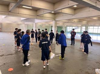 写真：青いジャージを着たスポーツ推進委員がモルックを教えています。その周りで生徒が熱心に話を聞いています