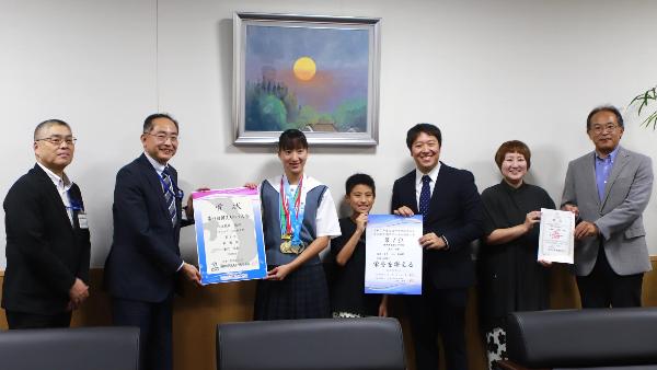 写真：歌代さんが3つのメダルを首から下げて賞状をもつ歌代選手。副市長・教育長、訪問者らと一緒に記念のカメラに収まりました。