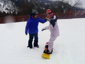 写真：参加者が講師に支えられながら斜面をスノーボードを履いて滑っています