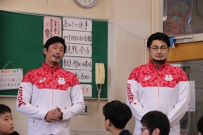 赤いジャージを着た男性2人が小学校で3人の子供たちに話をしている写真