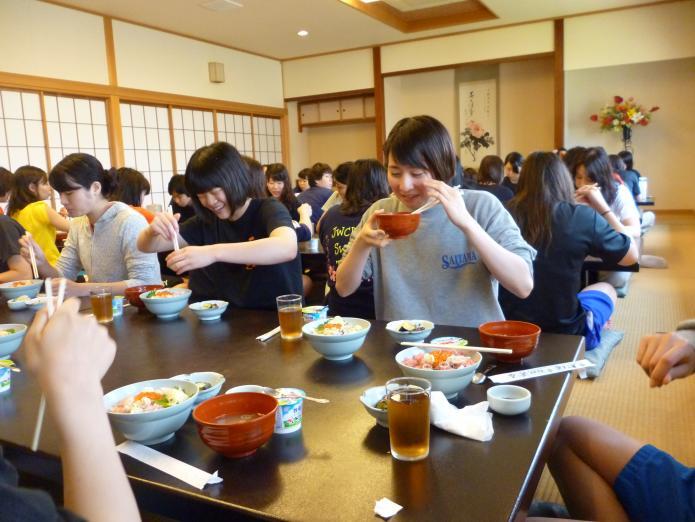 広い和室で3列の食卓に並んでいる大勢の学生それぞれに用意された丼ものやみそ汁ヨーグルトを食べているの写真