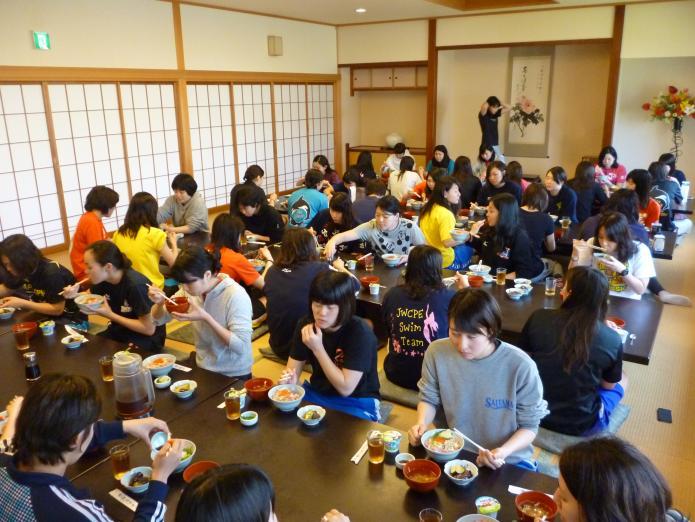 30名前後の大人数の女子大学生が旅館の広間でねぎとろ丼やみそ汁を食べている写真