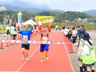 写真：男性2人がゴールした様子です。男性1人がビールくださいと書かれたタオルを頭の上に掲げ笑顔で走っています