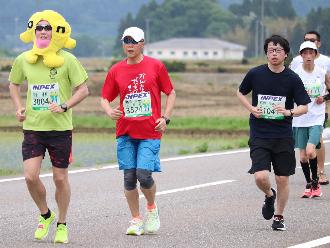 写真：ハーフマラソン折り返し付近の様子。ランナーが一生懸命走っています。写真左側の男性は着ぐるみをかぶり走っています