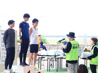 写真：表彰式の様子。男性3人が表彰台に立ち、実行委員長が表彰状を読み上げています