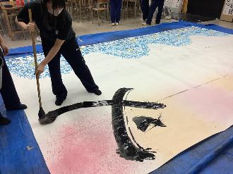 写真：高校生が大きな用紙に、「必勝」の「必」の3画目を書いています。使用する筆は高校生の肩ほどの長さです。