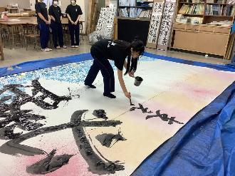 写真：「支え合った」と書く高校生。手には墨の入ったコップと太筆を持っています。