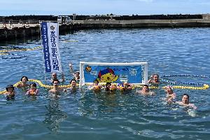 写真：水球体験会参加者の集合写真。漁港の中に設置した水球ゴールの前に参加者・関係者らが並んでいます。