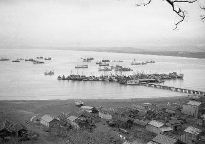 画像：砂浜から続く桟橋の奥に、ヨットや船などが停泊しています。モノクロ写真