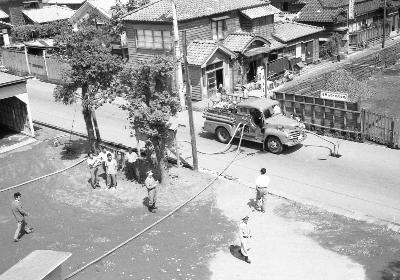 写真：トラック型の消防車から、放水用のホースが2本伸びています。周囲には訓練に参加する市民がいます。モノクロ写真