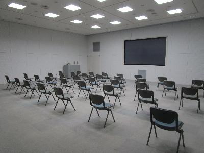 大型スクリーンの前に演台、テーブル付きの椅子がスクリーンに向かって並んでいる多目的ホールの写真