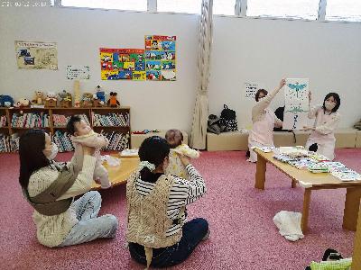 写真：お母さんの膝に抱っこされた赤ちゃんを前に、ブックスタートで渡す絵本を紹介しているところ