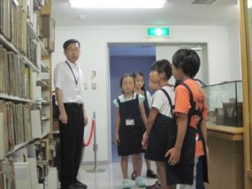 おそろいのエプロンを身に着けた小学生たちが、図書館司書から図書館の説明を受けている写真