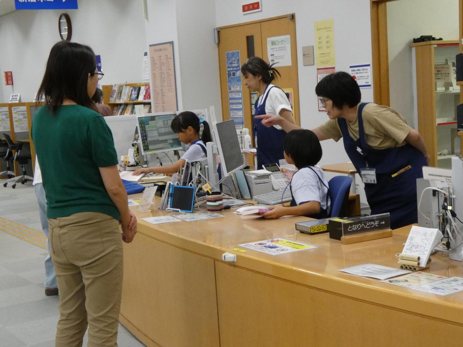 写真：職員が付き添いながらカウンターで資料の貸し出し・返却業務を行う2人の子ども