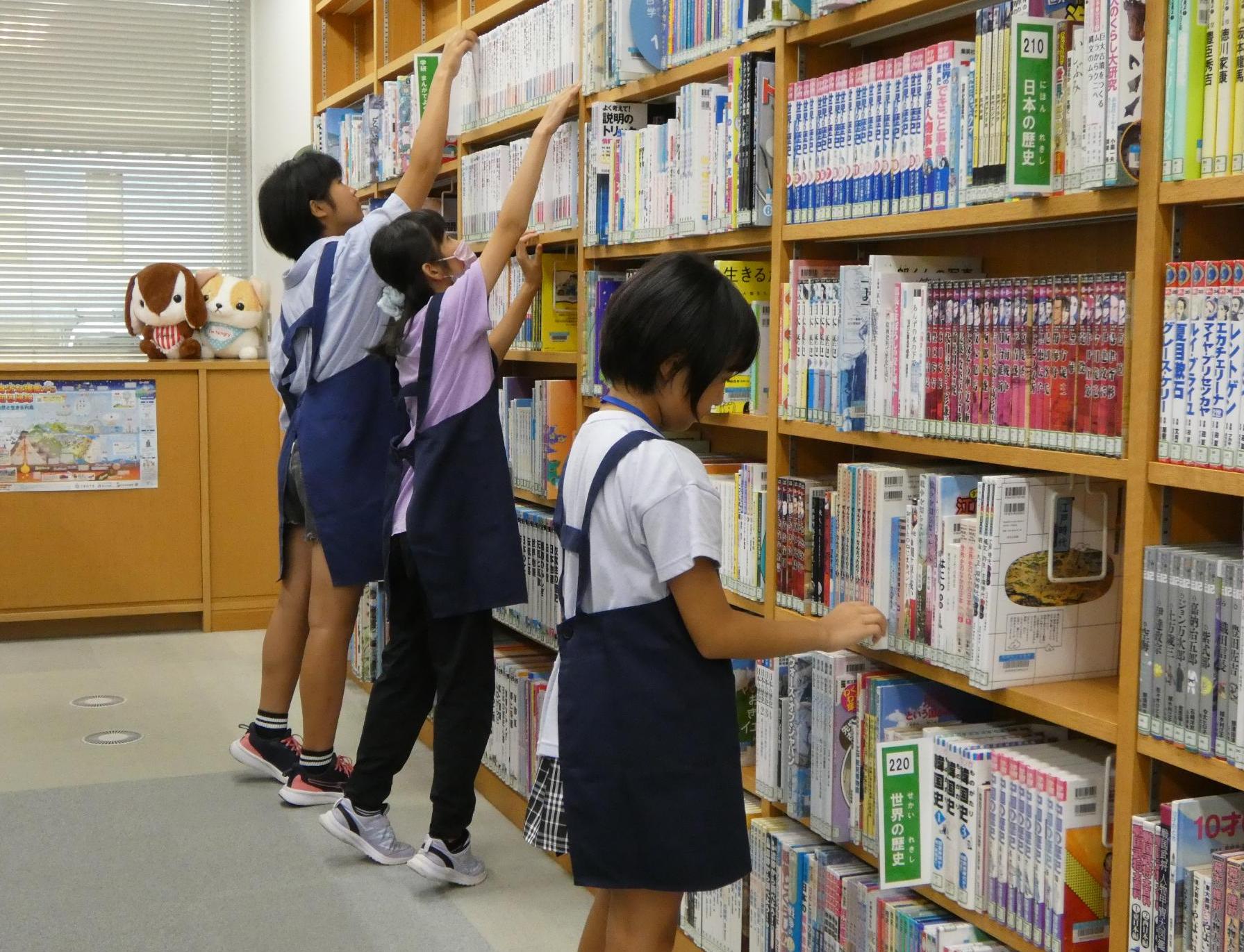 写真：3人の子どもが書架の整理をしています。奥の2人は高い棚に本を戻そうと背伸びをしています