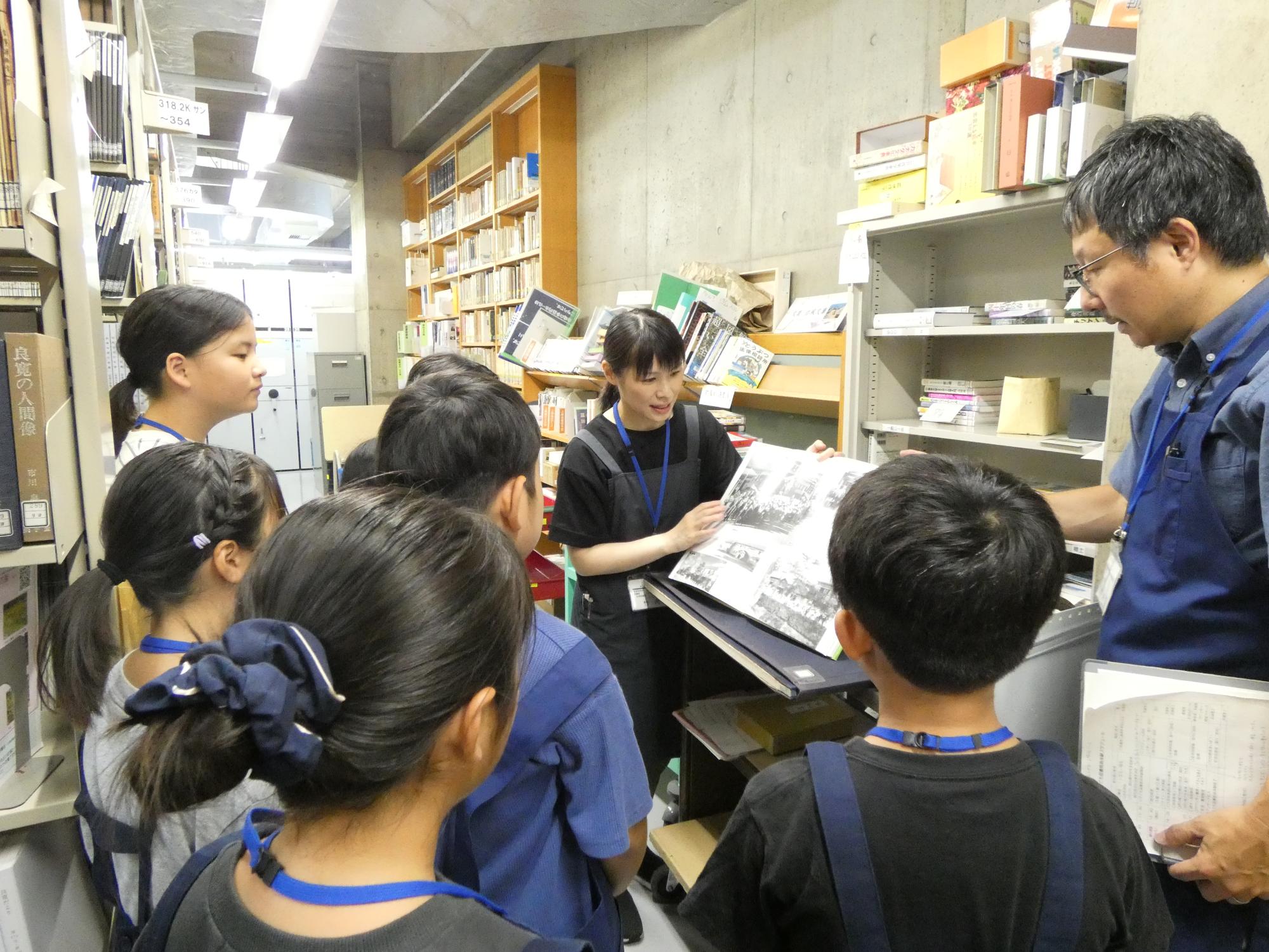 写真：1階書庫の見学の様子。保存された昔の新聞を見せながら、職員が子どもたちに説明しています。