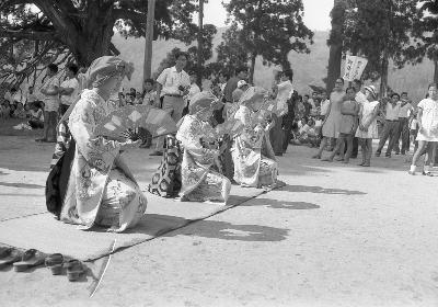写真：神社の境内にむしろを敷き、そのうえで綾子舞小歌踊を披露する3人の女性。頭に布を巻き、振り袖姿で扇を持っています。モノクロ写真