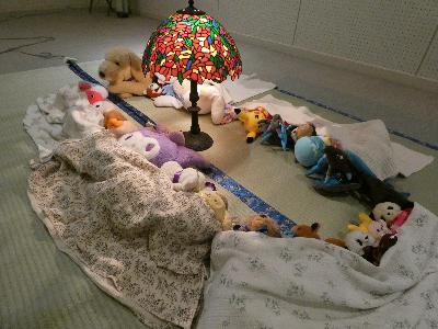 写真：ランプを囲んで、ぬいぐるみたちが毛布を掛けてお昼寝をしているところ