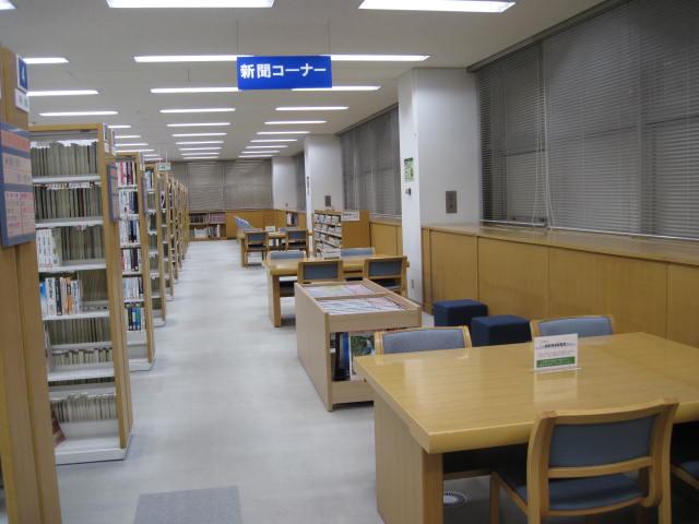 壁側にテーブルが置かれ、左側に本棚が並んでいる新聞コーナーの写真