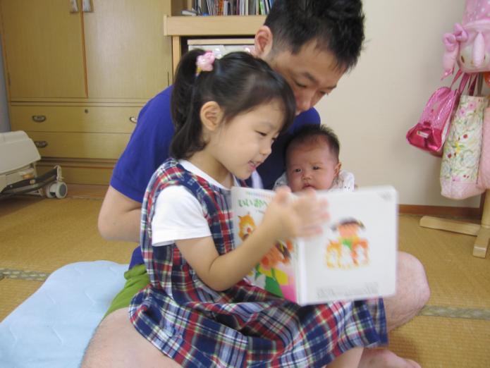 お父さんの膝の上で女の子と赤ちゃんが座り、女の子が赤ちゃんに絵本を読んでいる写真
