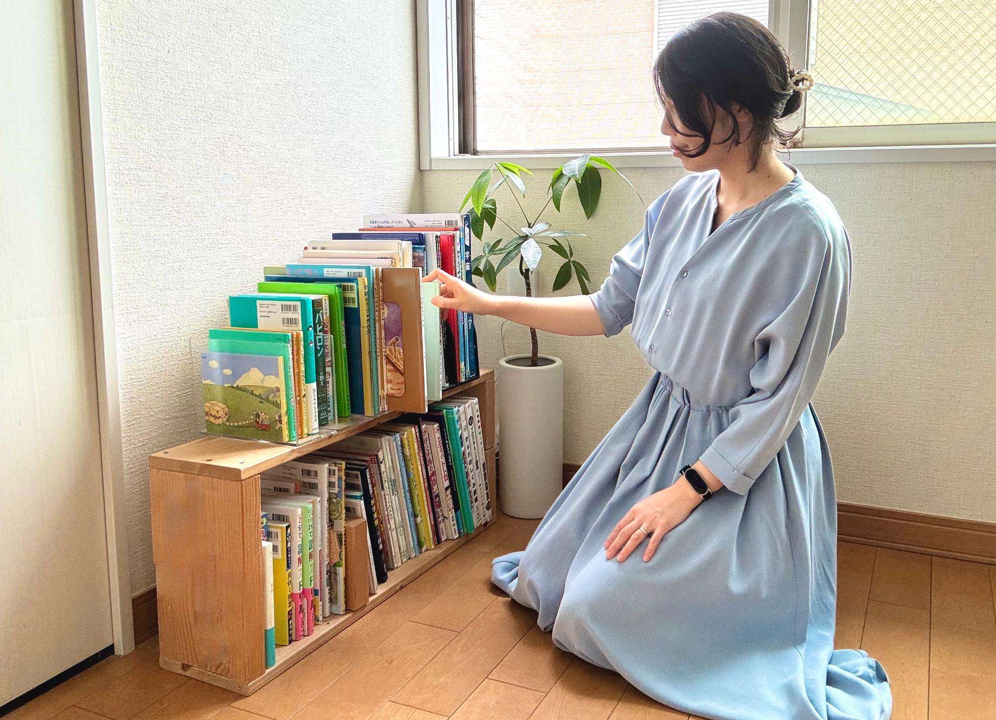 写真：整頓された本棚の前に膝をついた女性が、本棚から絵本を取り出そうとしています。