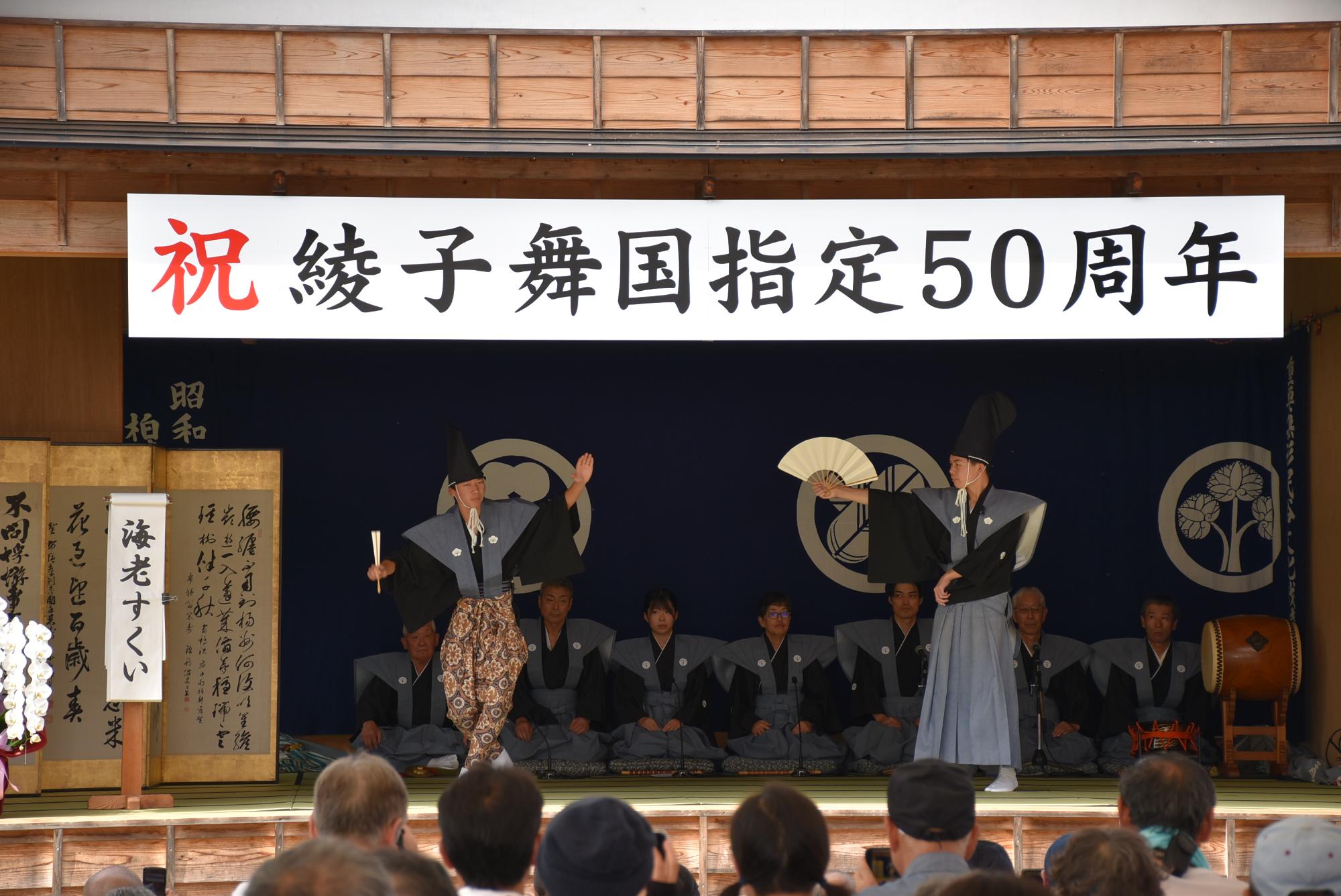 二人の男性が扇をもち、一人は中腰で、一人は偉そうに真っすぐ立って、ユーモラスに狂言を演じている写真