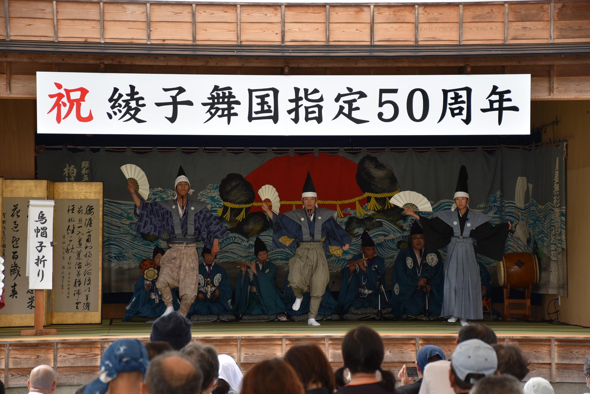 三人の男性が右手に持った扇を上げながら狂言を演じている写真