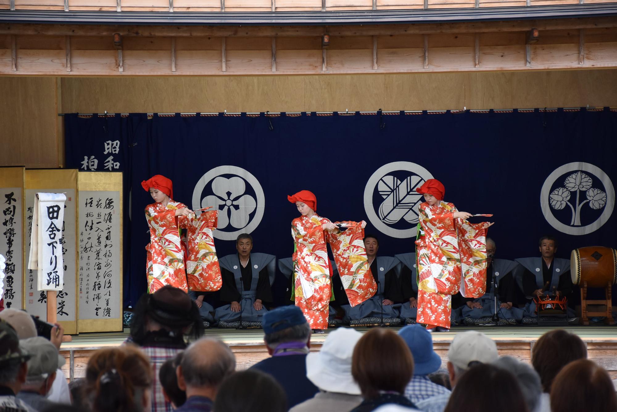 三人の赤く華やかな着物を着た女性が、扇を広げ、胸のあたりに水平に持ち踊っている写真