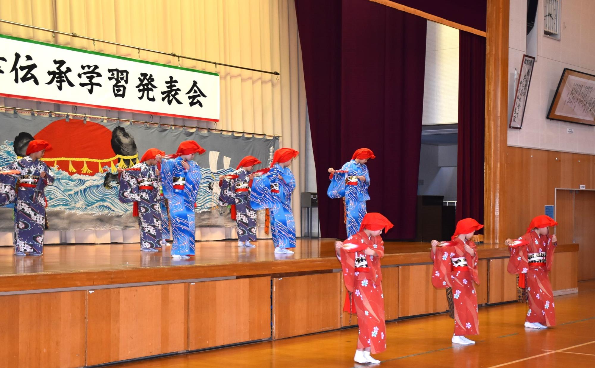 写真：9人の女子児童生徒がステージの上と下に分かれ、両手に扇子を持ち、腰を少し落とした状態で、顔を伏せながら舞っています。