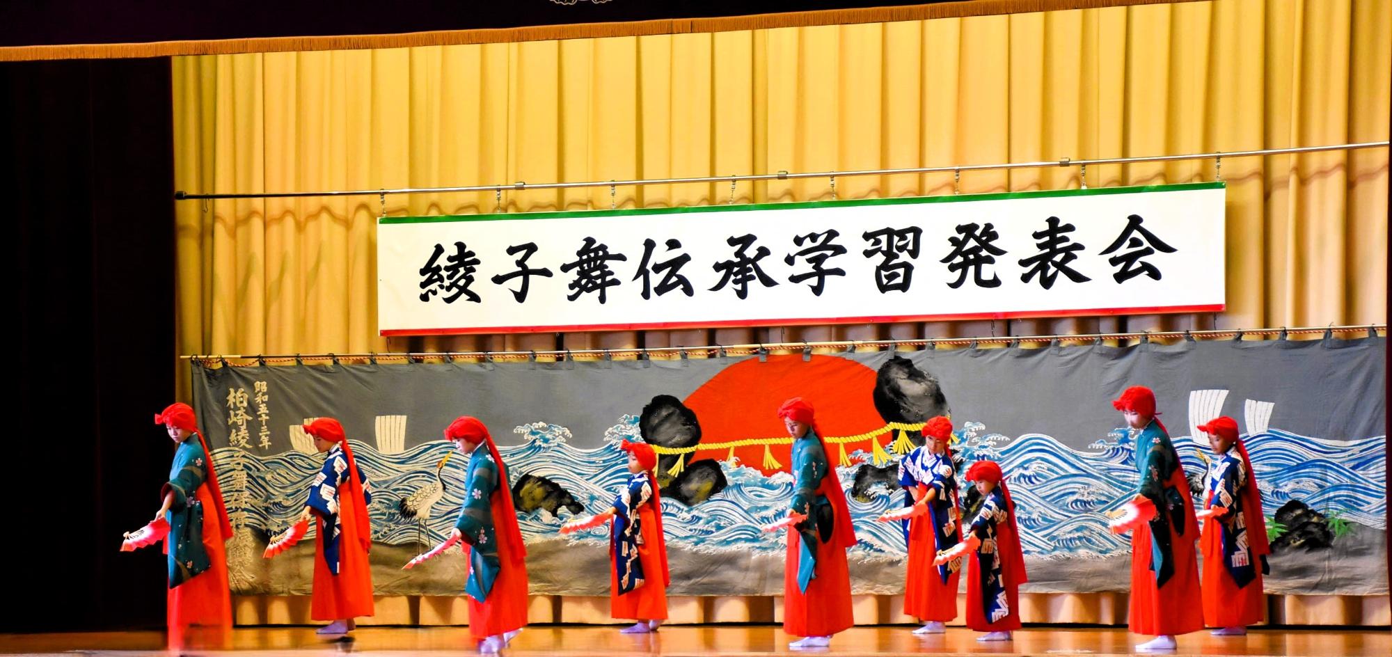 写真：赤い袴にユライを付けた9人の女子児童・生徒が、左手に持った扇子を前方下に伸ばし踊っているところ。