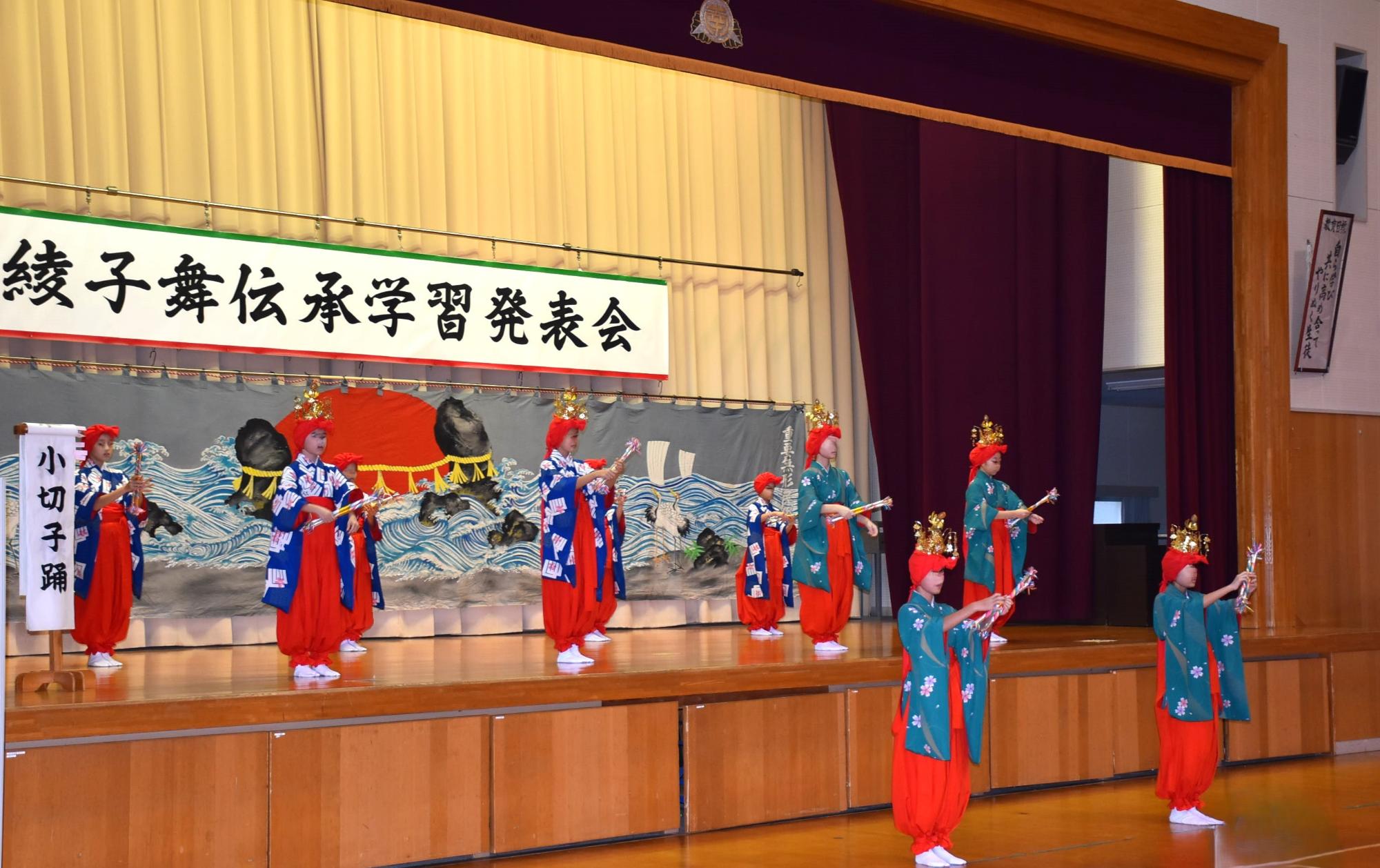 写真：10人以上の女子児童・生徒が、ステージ上と下に分かれて、踊っています。頭に天蓋(てんがい)をつけ、綾竹を自分の前で打ち鳴らしています