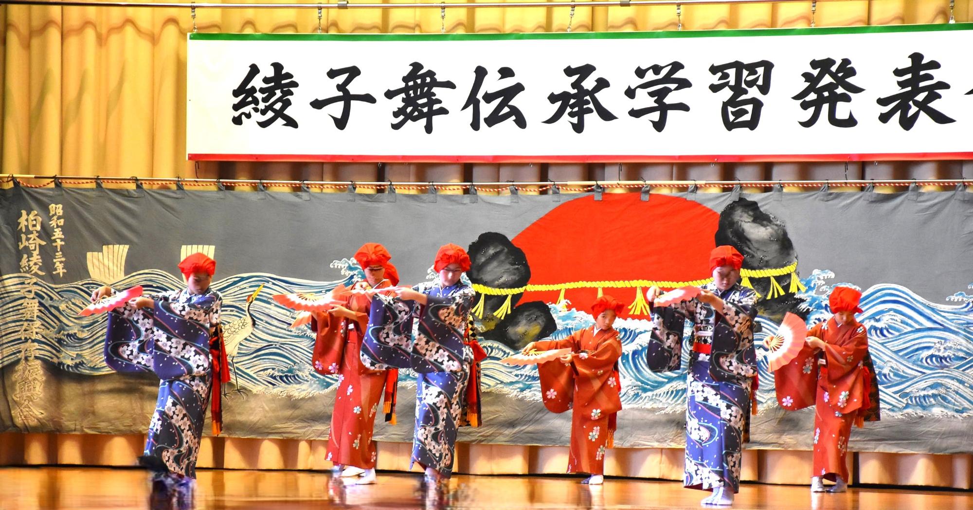 写真：赤いユライを頭に被り、7人の女子児童・生徒が、少し腰を落として両手で持った扇を体の右側に移動させながら舞っています