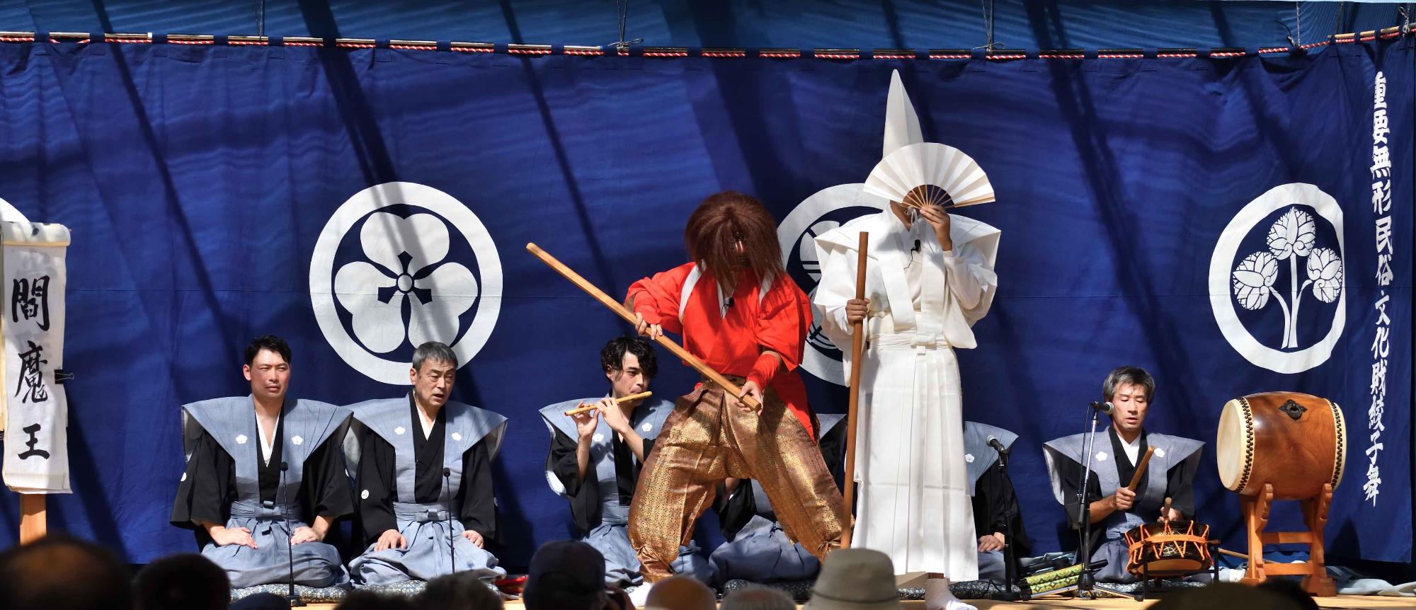赤色の着物を着た男性と白色の着物を着た男性が二人狂言を演じている写真