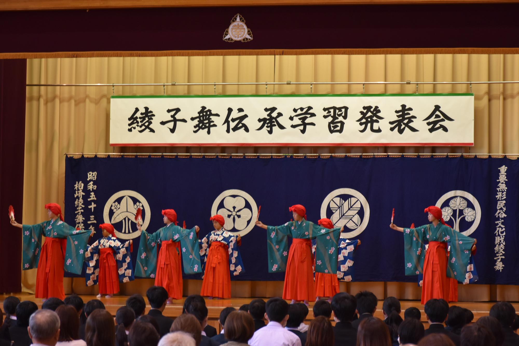 写真：8人ほどの女子児童・生徒が右手に扇を持ち、両手を広げ舞っているところ。ユライと呼ばれる赤い布を頭に巻き、振り袖に緋色の袴をはいています。