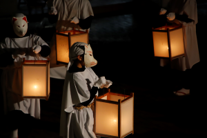 写真：灯りのともった行燈を両手で持ち、狐のお面に白装束姿の人たちが、かがり火の周りを列になって歩いているところ