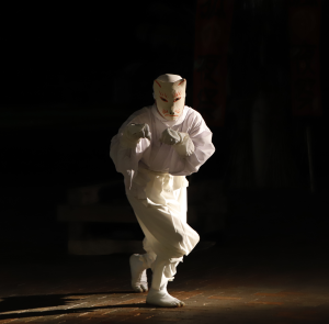 写真：腰をひねり、腕を前にして手首を下に曲げ、こぶしを軽く握って立つ白装束とお面で狐に扮した人が光に照らし出されています。